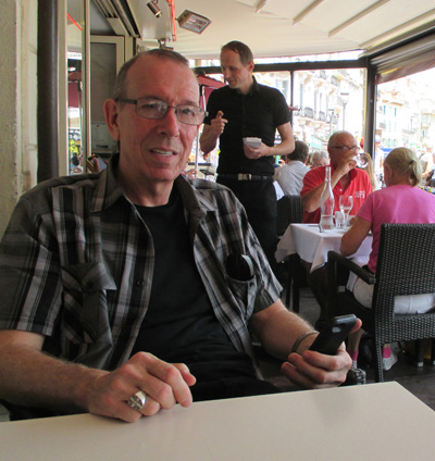 photo shows Gene at a small table under an awning, smiling and holding his smartphone.